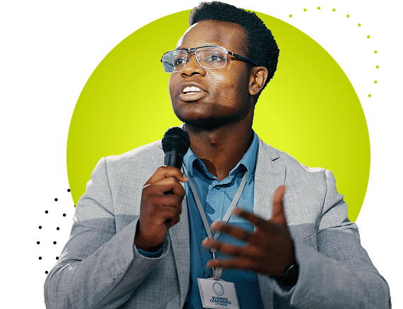 Man in a suit jacket and collared shirt holding a microphone mid-speech, wearing eyeglasses and a lanyard badge